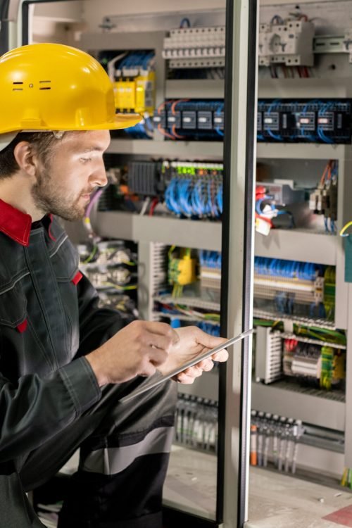Contemporary young engineer in workwear and hardhat scrolling through data in tablet while squatting by one of industrial machines